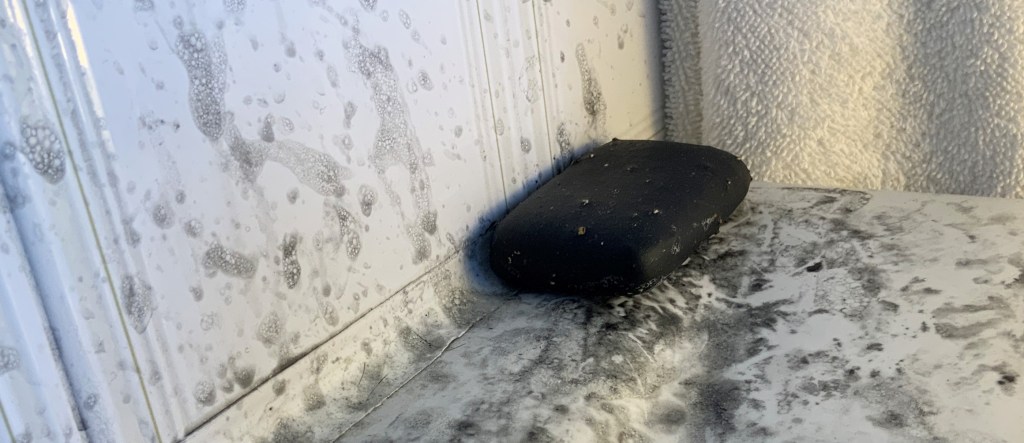 This image shows a bar of Kaolin and Charcoal soap on the corner of a white bath.  Behind the bath is a white towel.  The bath and white bath tiles are covered with grey/black splash marks and bubbles.