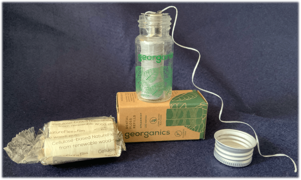 Blue background with a slim glass jar containing a reel of plant based dental floss, an aluminium screw top lid, cardboard box and two replacement reels in a cellulose based clear package