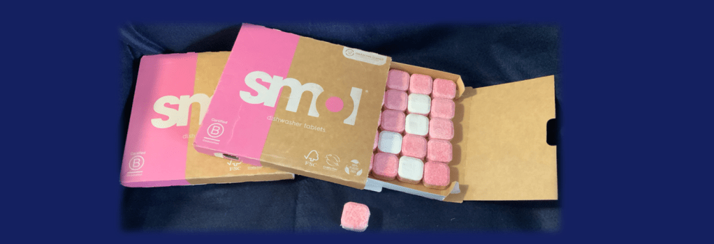 Image, blue background with a photo of two packs of Smol dishwasher tablets.   One pack is open showing the pink and white tablets which are wrapped in a plastic-free soluble clear film.